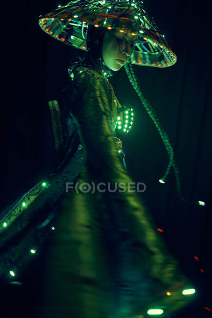 Frau in authentischem Outfit und vietnamesischem Hut mit leuchtender Beleuchtung steht während des Auftritts im dunklen modernen Studio auf schwarzem Hintergrund — Stockfoto
