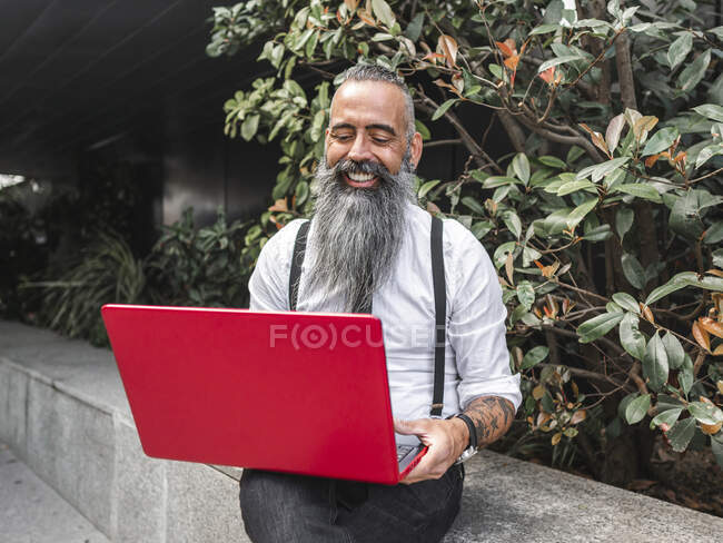 Positivo hipster maschio freelance in abbigliamento formale navigazione netbook moderno mentre seduto sul bordo di pietra vicino a piante verdi per strada durante il lavoro online — Foto stock