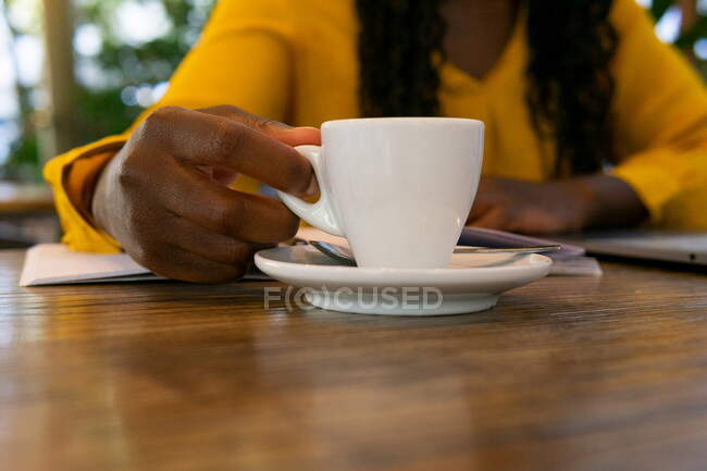 Crop anonymous African American person with white cup of hot drink sitting at wooden table in modern cafeteria on blurred background — Stock Photo