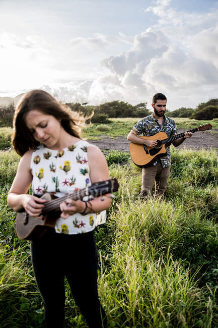 Спокійна жінка в повсякденному одязі грає на укулеле і стоїть в зеленому полі проти концентрованого друга, грає на гітарі в природі в сонячний день — стокове фото