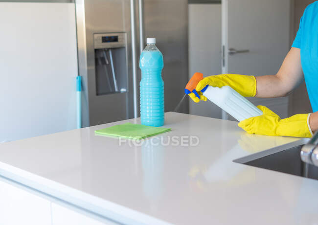 Обрізати анонімну жінку в жовтих гумових рукавичках, розпилюючи миючий засіб на тканину під час прибирання світлої кухні — стокове фото
