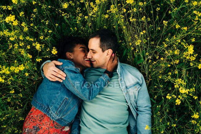 Loving young multiracial couple in casual clothes kissing while lying in lush blooming meadow during romantic date on sunny day — Stock Photo
