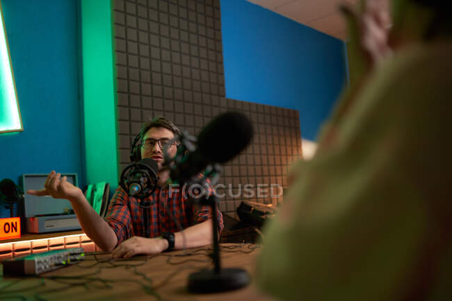 Young male radio host in checkered shirt and headphones sitting at table with microphone and communicating with crop anonymous colleague during podcast recording in studio — Stock Photo