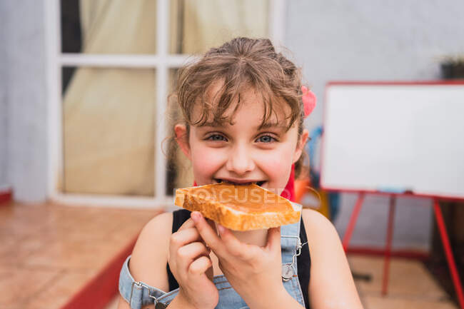 Симпатичная девушка в джинсе в целом глядя на камеру во время еды свежий хлеб со сладким вареньем в светлом помещении дома — стоковое фото