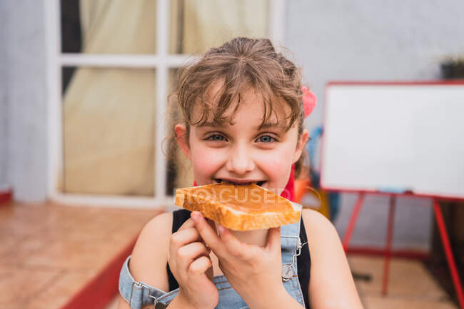 Cute girl in denim overall looking at camera while eating fresh bread with sweet jam in light room at home — Stock Photo