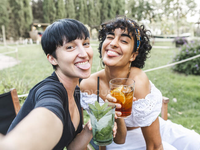 Delighted multiracial best female friends with glasses of cocktails chilling in summer park and taking selfie on smartphone while sticking out tongue — Stock Photo
