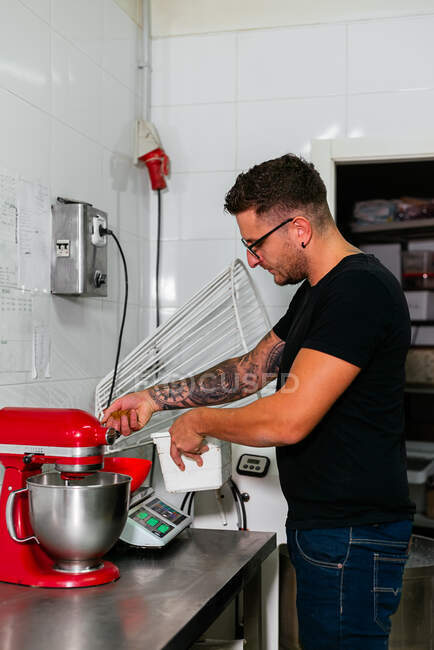 Seitenansicht eines jungen tätowierten Konditors in lässiger Kleidung und Brille, der Mehl auf elektronischer Waage wiegt, steht am Tisch mit Mixer in der Küche — Stockfoto