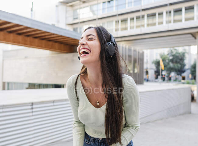 Mujer alegre con el pelo castaño largo en ropa casual y auriculares escuchando música y riendo en la calle de la ciudad a la luz del día - foto de stock