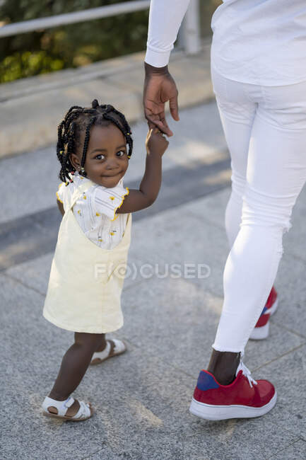Щаслива афроамериканська дівчинка у світлому одязі, тримаючи за руки невпізнаного батька під час прогулянки по вулиці в сонячний день. — стокове фото