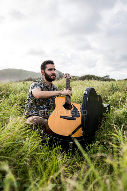 Vista lateral de calma músico masculino em roupas casuais sentado na grama verde e abertura caixa preta de guitarra acústica na costa perto do mar à luz do dia — Fotografia de Stock