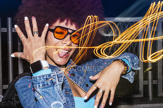 Crop of cheerful female with sunglasses looking at camera while standing near fence with light effect in evening time and sticking out his tongue while moving his hands — Stock Photo