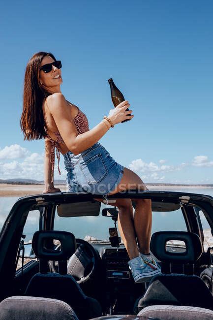 Full body of happy young female in summer outfit and sunglasses holding beer while sitting on roof of safari car on shore of river — Stock Photo