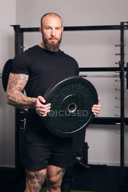 Powerful sportsman in sportswear standing with black weight plate in hands and looking at camera during workout in gym — Stock Photo