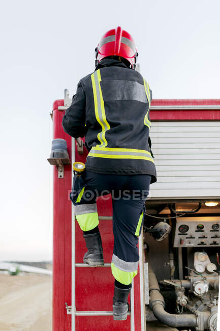 Back view of anonymous firefighter wearing red protective helmet and uniform standing on fire truck ladder and looking into the distance in daytime — Stock Photo