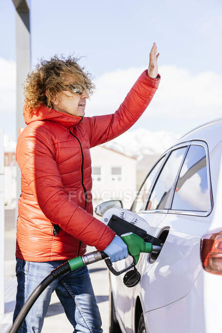 Side view of serious male with curly hair in outwear and latex gloves standing and waving hand while refueling car at gas station in sunny day — Stock Photo