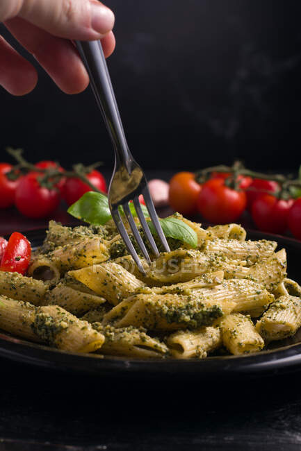Crop anonymous person with fork eating delicious pasta with green pesto sauce served on plate on black background — Stock Photo