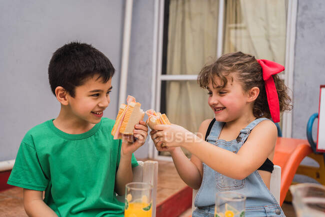 Niños positivos en ropa casual con sándwiches frescos en las manos sentados en sillas cerca de los vasos en la sala de luz en casa - foto de stock