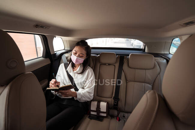 Mujer vestida formalmente con máscara protectora escribiendo en bloc de notas mientras monta en el asiento trasero del pasajero en cabina cómoda - foto de stock