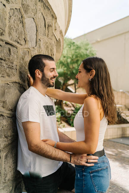 Visão lateral da jovem mulher étnica positiva em roupas casuais abraçando o namorado barbudo sorridente apoiando-se no prédio de pedra envelhecido no parque da cidade no dia ensolarado — Fotografia de Stock
