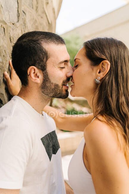 Vista laterale della giovane coppia etnica innamorata in abiti casual che si abbracciano e si baciano in piedi vicino al muro di pietra sulla strada della città — Foto stock