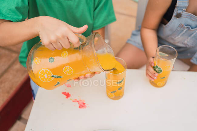 Menino anônimo derramando suco doce de jarro enquanto sentado perto da menina à mesa com sanduíches frescos na placa na sala de luz — Fotografia de Stock