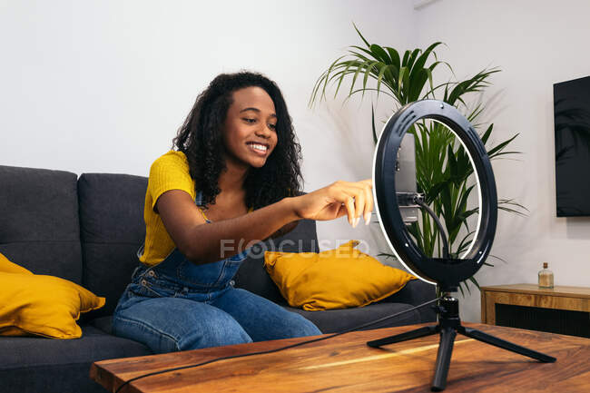 African American woman in denim clothes smiling while sitting on sofa and touching smartphone on glowing ring lamp — Stock Photo