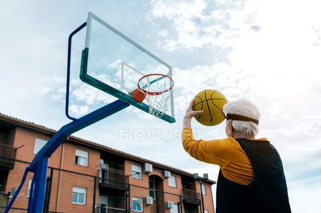 Back view of anonymous mature female in activewear throwing ball into hoop while playing basketball on public sports ground on street — Stock Photo