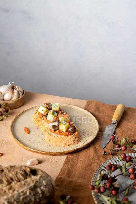 Tostadas con cubos de queso y rebanadas de champiñones servidos en plato cerca de pan fresco y tazón de ajo en la cocina - foto de stock