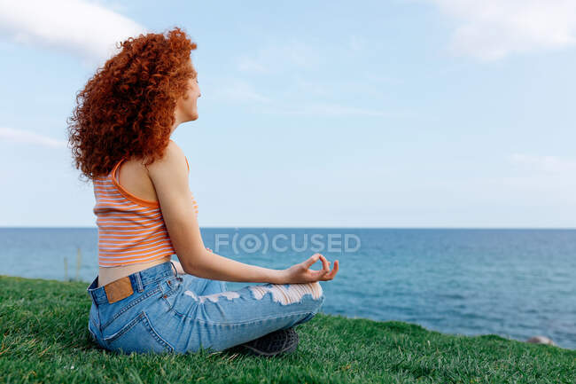 Vista laterale di donna spensierata con lunghi capelli rossicci in posa Padmasana con gesto zen sulla collina erbosa sulla riva del mare — Foto stock