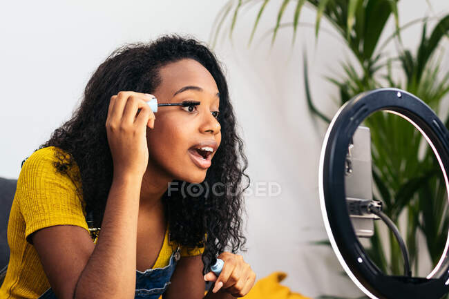 Чорна жінка наносить туш на вії, використовуючи передню камеру смартфона на блискучу кільцеву лампу — стокове фото