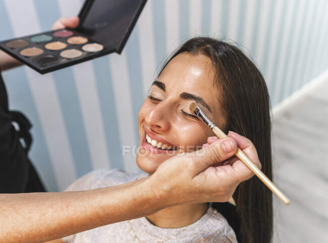 From above of crop visagiste applying makeup with brush on eyelid of young smiling female — Stock Photo