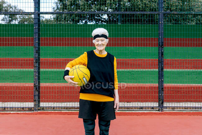 Femme mature positive en vêtements de sport et bandeau regardant la caméra tout en se tenant avec la balle pendant le match de basket-ball — Photo de stock