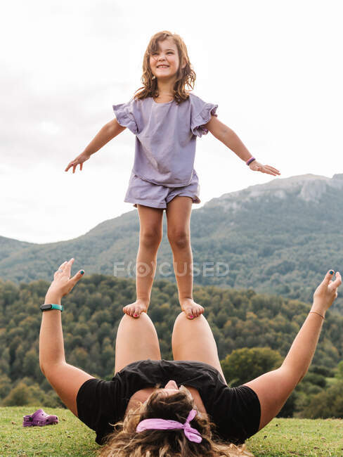 Positives Mädchen balanciert und steht auf Knien einer gesichtslosen Mutter auf einer Rasenfläche vor bergigem Gelände mit grünen Bäumen in der Natur — Stockfoto