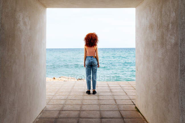 Обратный вид на всю длину неузнаваемой женщины-туристки, любующейся волнистым морем с бетонного прохода на побережье — стоковое фото