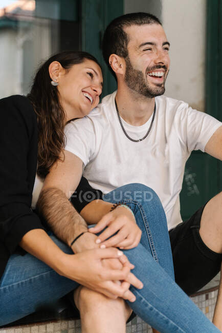 Jovem mulher étnica alegre em roupas casuais abraçando namorado enquanto rindo juntos sentados nas escadas na rua — Fotografia de Stock