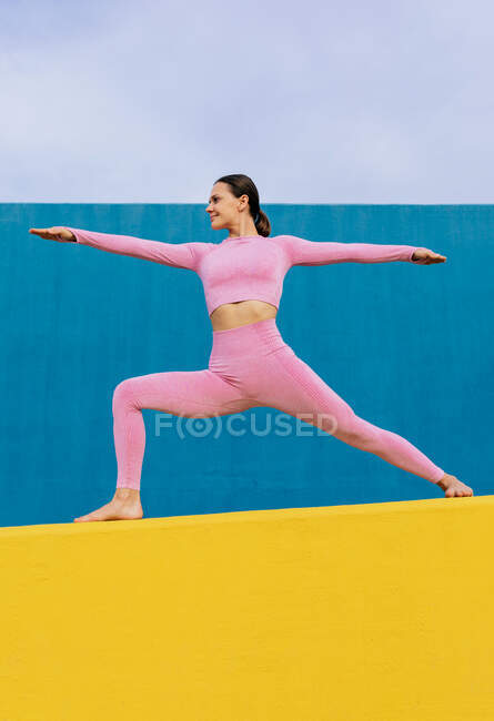 Low angle of cheerful flexible female standing in Virabhadrasana and doing yoga on vibrant yellow and blue background — Stock Photo