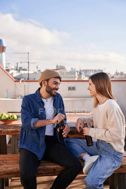 Compañeros de piso positivos jóvenes masculinos y femeninos sentados en el banco con botellas de cerveza y tener una conversación agradable en la terraza - foto de stock