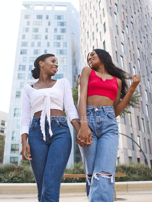 From below of optimistic African American lesbians in stylish clothes looking at each other and holding hands while walking on street with buildings — Stock Photo