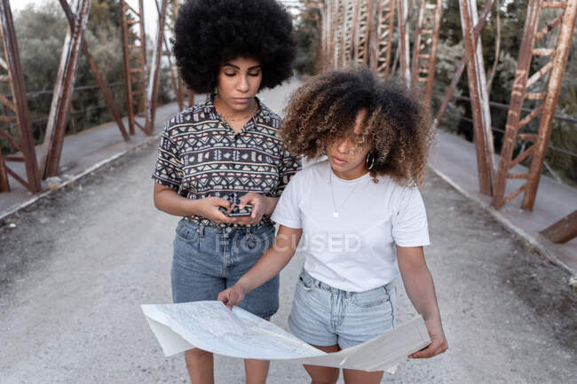 Amigas afro-americanas sérias em pé na ponte velha e em busca de direção no mapa de papel e no celular — Fotografia de Stock