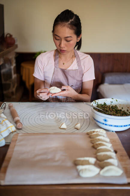 Mujer en ropa casual y delantal relleno albóndigas con carne mientras se prepara jiaozi tradicional chino en la cocina - foto de stock
