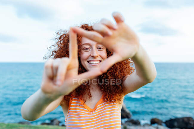 Positives Weibchen mit langem, lockigem Ingwerhaar, das Dreieck an der Küste mit Felsbrocken formt und in die Kamera blickt — Stockfoto