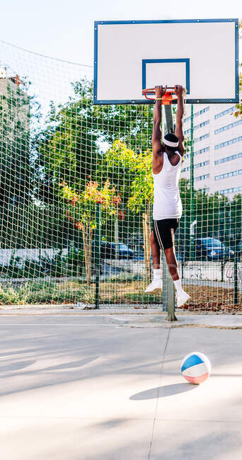 Vue arrière du joueur afro-américain anonyme de basket-ball suspendu à un cerceau métallique sur une aire de jeux en été — Photo de stock