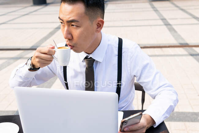 Jeune entrepreneur asiatique poignant avec tasse de boisson chaude et netbook regardant loin à la table de cafétéria urbaine en plein jour — Photo de stock