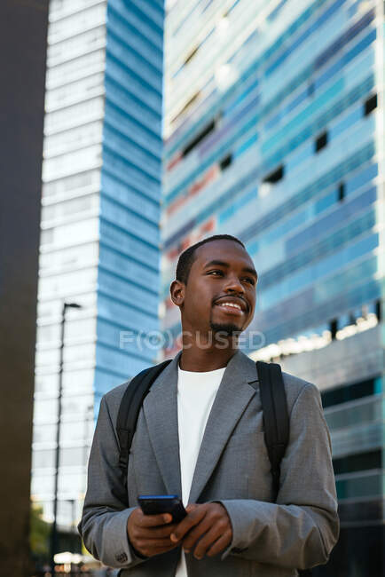 Smiling African American businessman in formal wear with backpack browsing mobile phone in downtown and looking away with toothy smile — Stock Photo