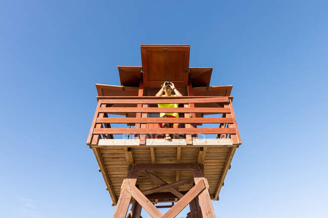 Bajo ángulo de mirar a través de binoculares en la torre de vigilancia de madera mientras supervisa la seguridad en el mar contra el cielo azul sin nubes - foto de stock
