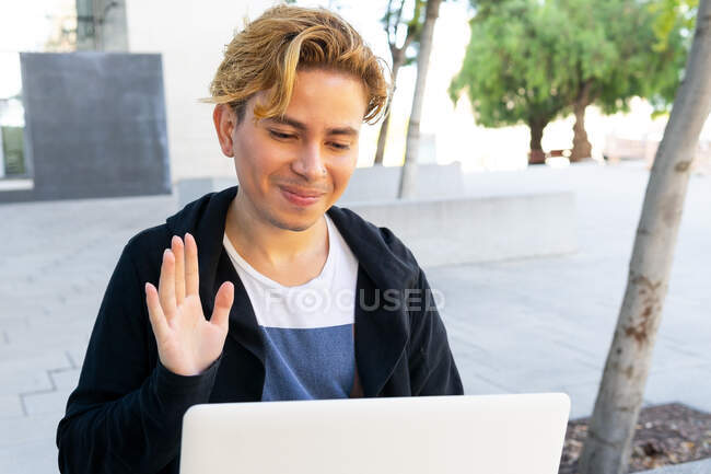 Cheerful young male in casual outfit waving hand while having video chat on modern netbook on street of city with trees — Stock Photo