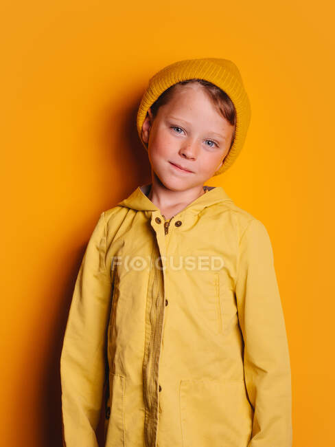 Happy boy in vivid yellow raincoat jacket and beanie hat laughing and looking at camera against yellow background in studio — Stock Photo