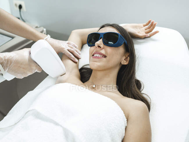 De acima mencionado mestre jovem em óculos fazendo o procedimento de remoção de cabelo a laser com equipamento profissional na mão no salão de beleza leve — Fotografia de Stock