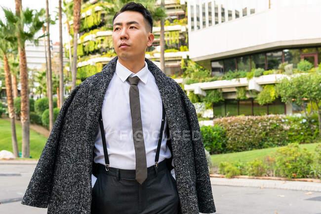 Bem vestido jovem empresário asiático em gravata olhando embora enquanto passeia na estrada contra edifícios modernos na cidade — Fotografia de Stock