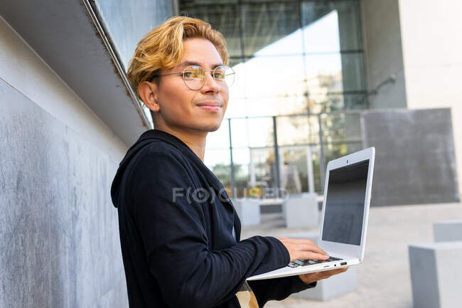 Joven freelancer masculino escribiendo en netbook moderno mientras está parado en la calle en la ciudad durante el trabajo en línea - foto de stock
